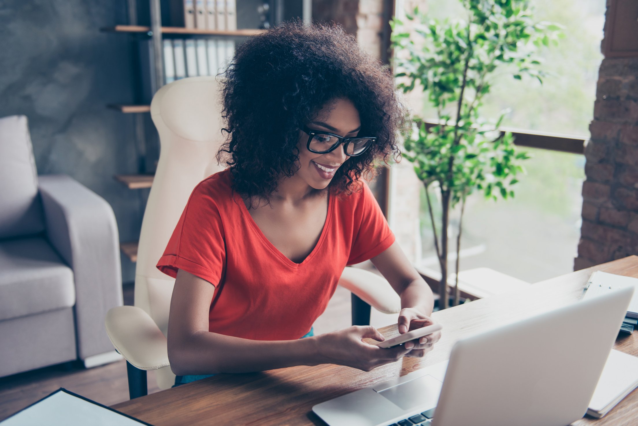 Portrait of cheerful trendy secretary sitting at desktop on armchair holding smart phone in hands using 5G wi-fi internet chatting with friends checking email searching contact enjoying free time